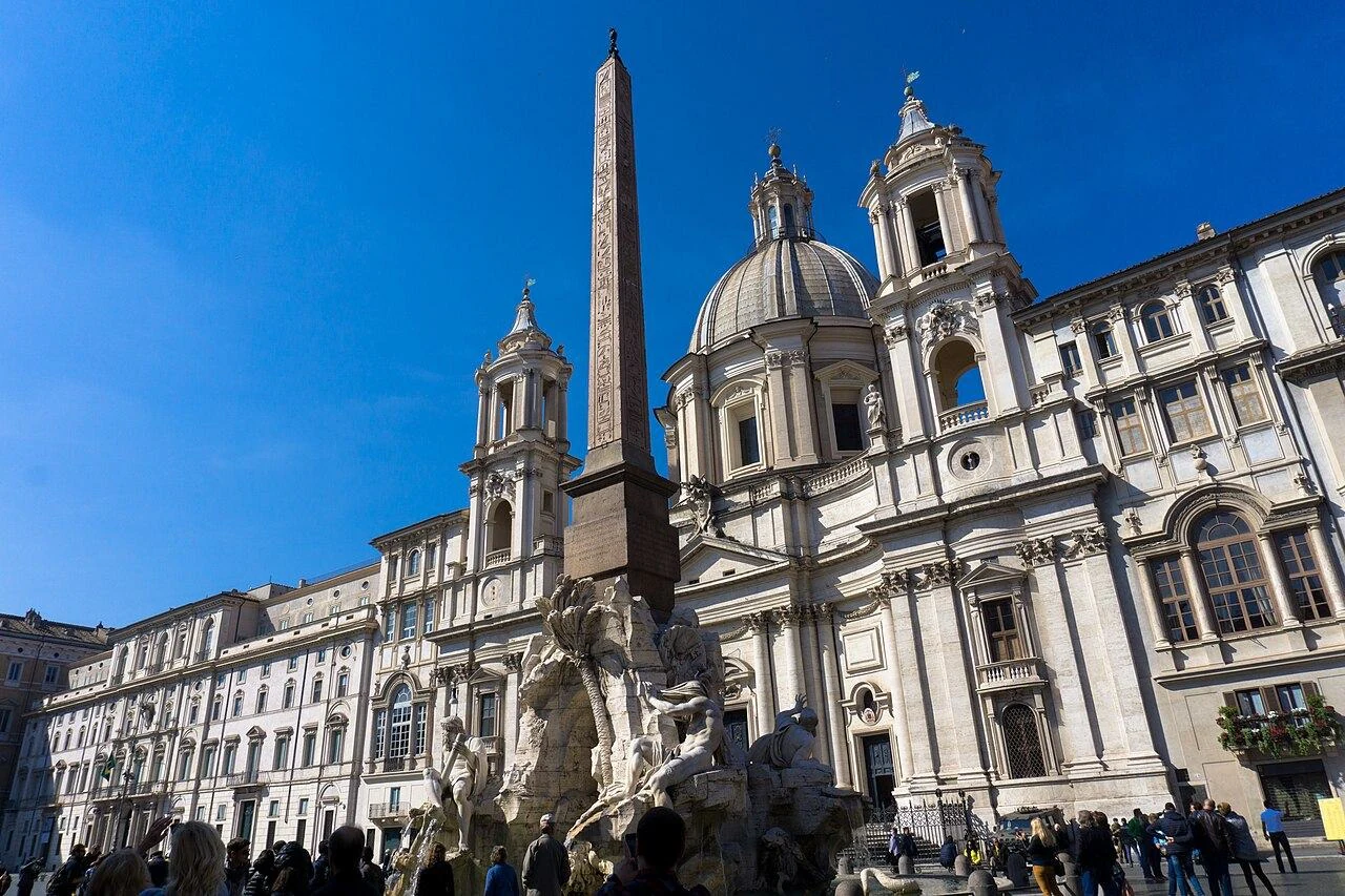 Vista diurna mais próxima da praça, com fonte, obelisco e Sant&rsquo;Agnese formando o conjunto clássico. Ela funciona como a 