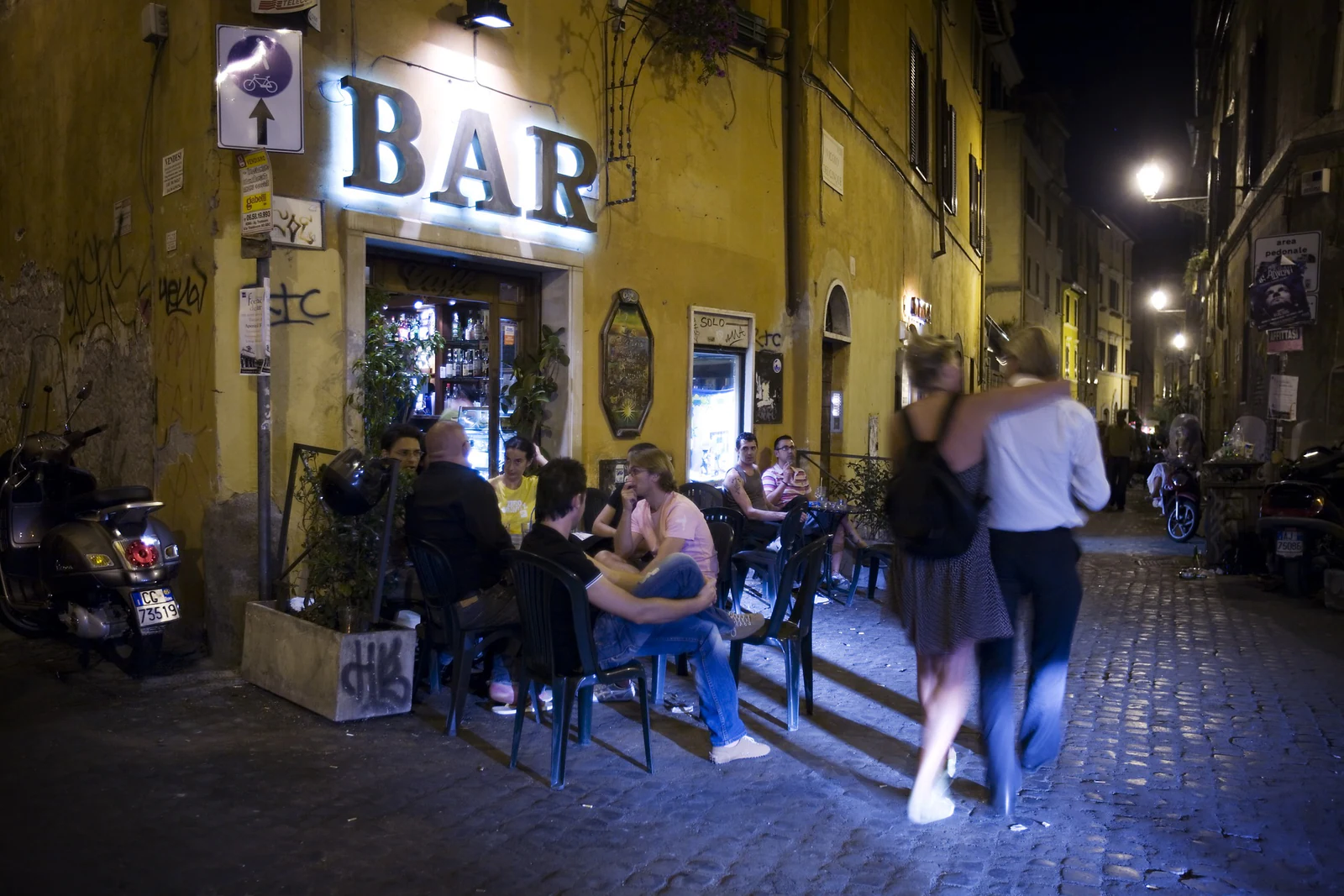 Uma rua de Trastevere à noite, com mesas, bares e pedestres ocupando o espaço. Esta imagem representa melhor o clima rea