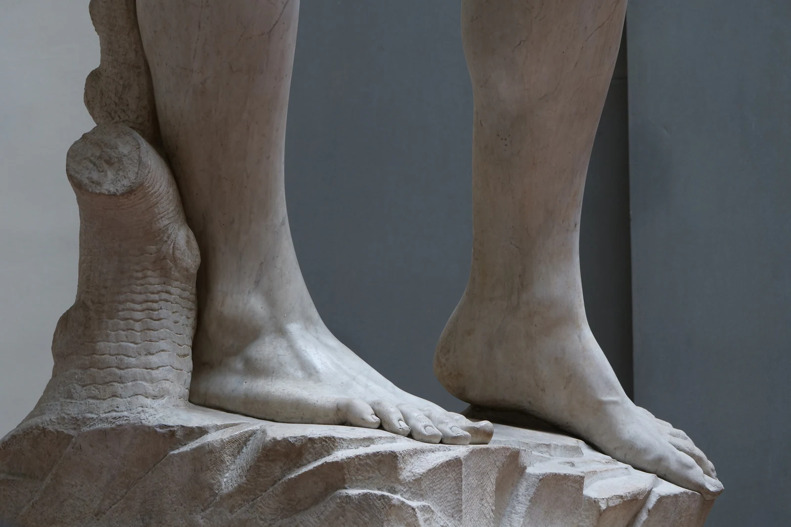 Pés, tornozelos e base vistos de perto. É uma boa foto para lembrar de olhar além do rosto: a escultura inteira é constr