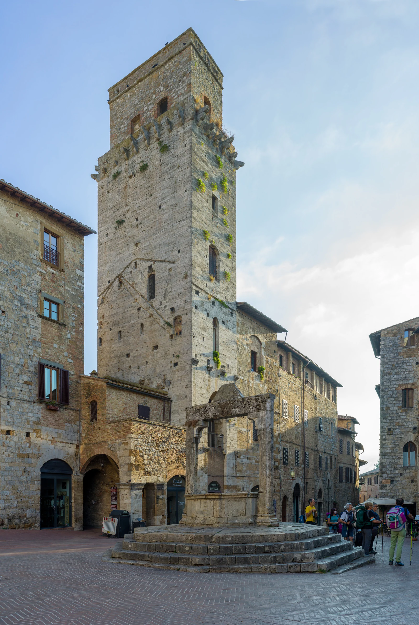 Piazza della Cisterna em uma vista vertical e muito aberta. O poço no centro, o piso inclinado e as fachadas medievais f
