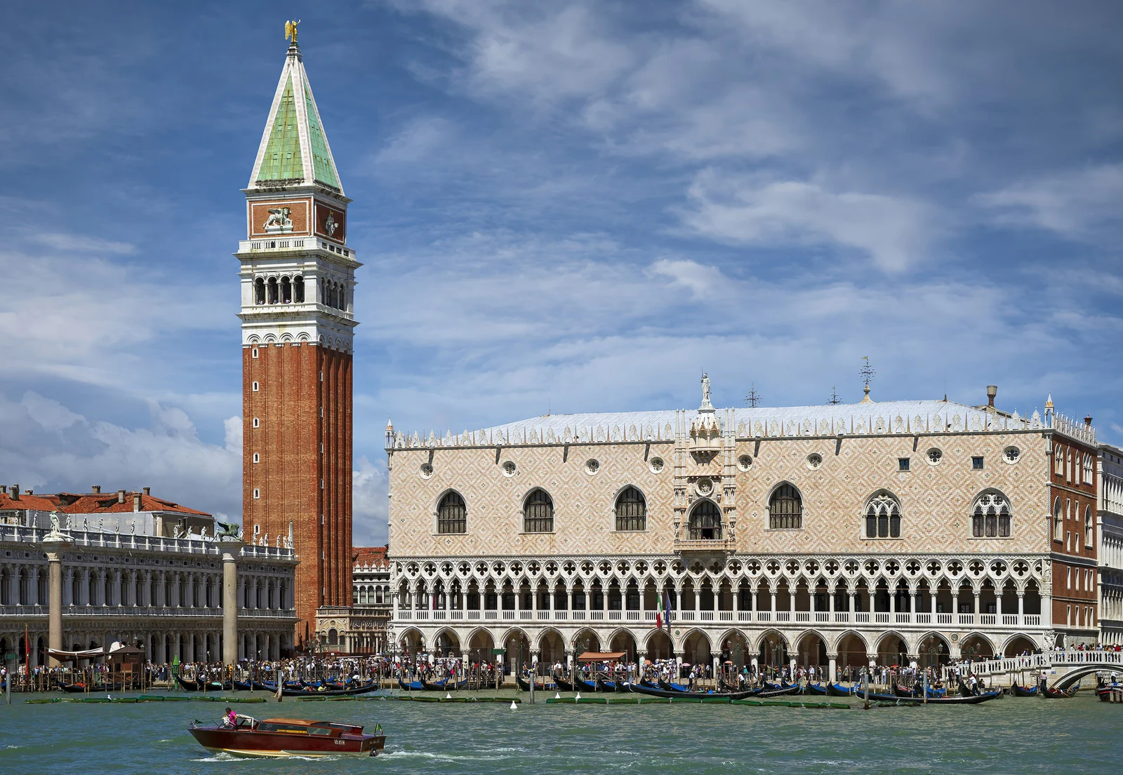 A fachada voltada para a laguna, com o Campanile e a Piazzetta compondo o centro político de Veneza. Repare na inversão que torna o palácio tão estranho e leve: duas ordens de arcadas abertas sustentam, visualmente, o grande volume fechado de mármore branco e rosado — como se a massa mais pesada estivesse flutuando.