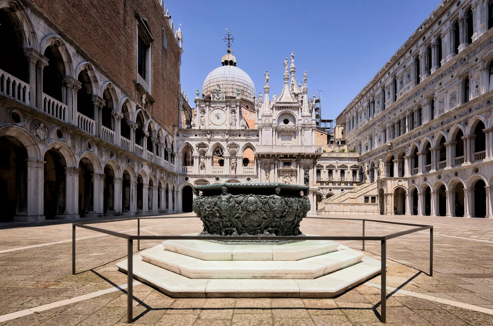 O pátio interno inteiro, com uma das cisternas de bronze em primeiro plano, a Scala dei Giganti ao fundo e San Marco surgindo por trás. É aqui que a visita muda de escala: o exterior parece renda gótica; por dentro, cada eixo conduz a uma escada, magistratura ou ritual de poder.