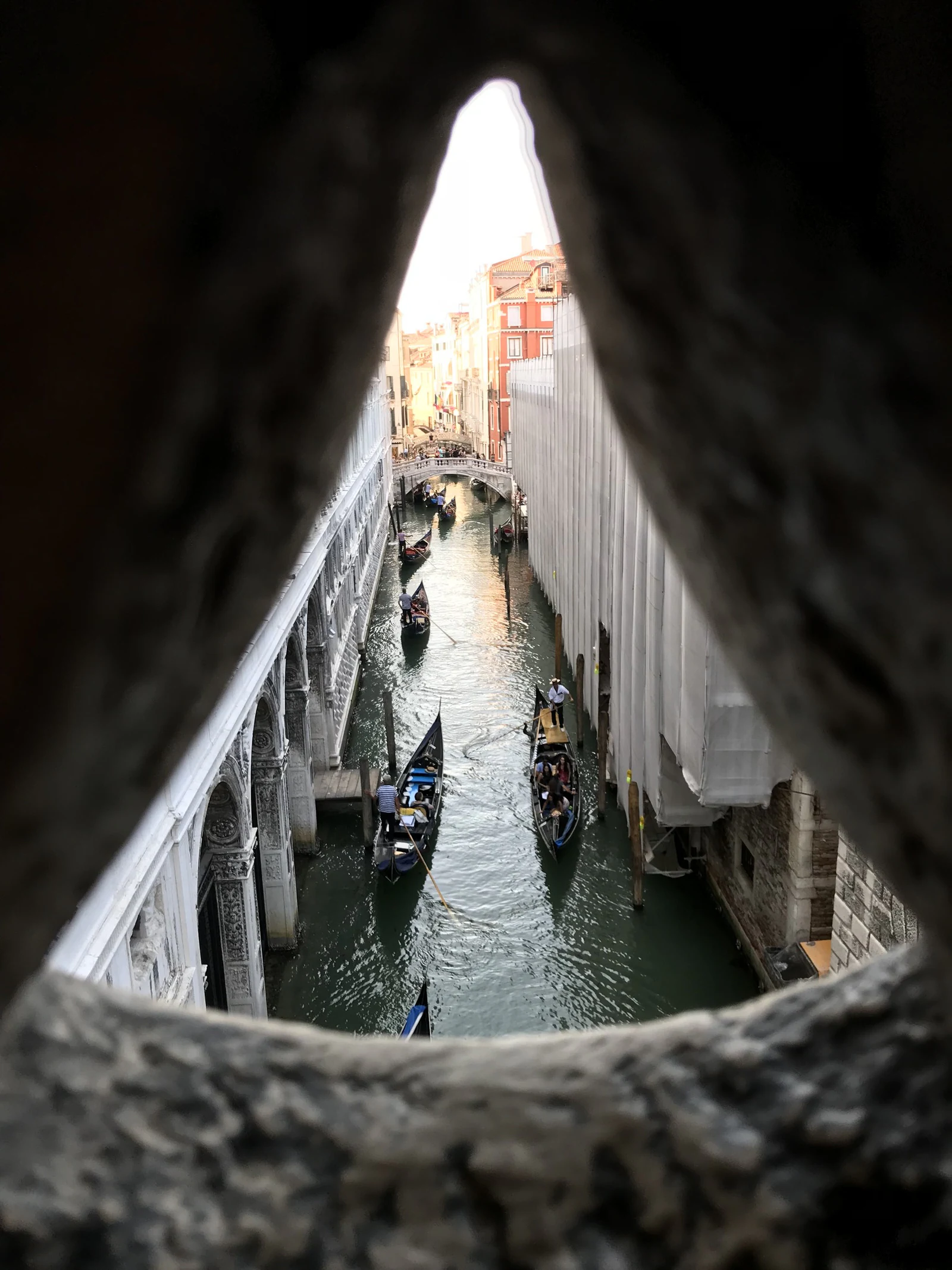 A vista real através da pequena grade de pedra da Ponte dos Suspiros. É muito mais estreita e fragmentada do que o mito sugere; procure San Giorgio e o movimento do canal entre os recortes. O nome e a história do ‘último olhar para Veneza&rsquo; foram popularizados no período romântico, séculos depois da construção da ponte.