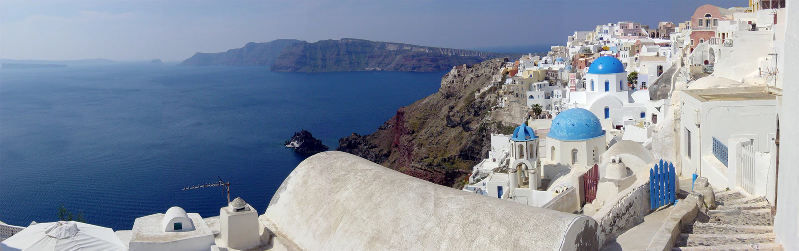 Este panorama explica a implantação: Oia ocupa uma faixa estreita no alto da parede vulcânica, enquanto as ilhas da caldeira ficam no horizonte.