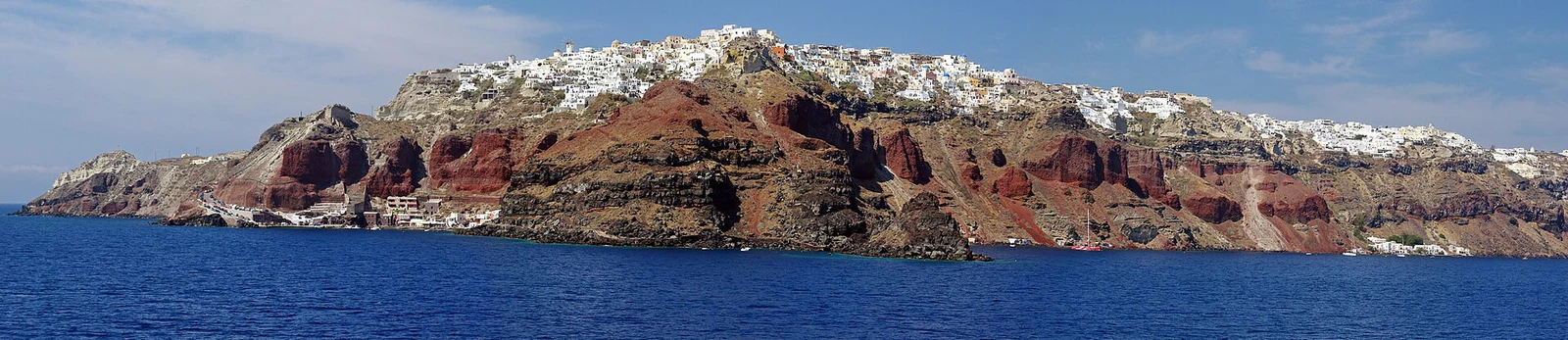 Oia vista do mar parece uma linha branca pousada sobre materiais vermelhos, ocres e negros.